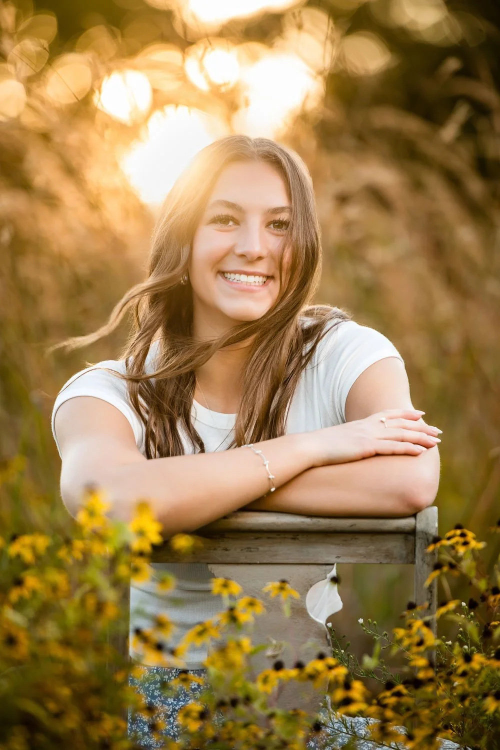 A young woman with long brown hair smiling at the camera, resting her arms on a wooden surface, in a field of yellow flowers during sunset