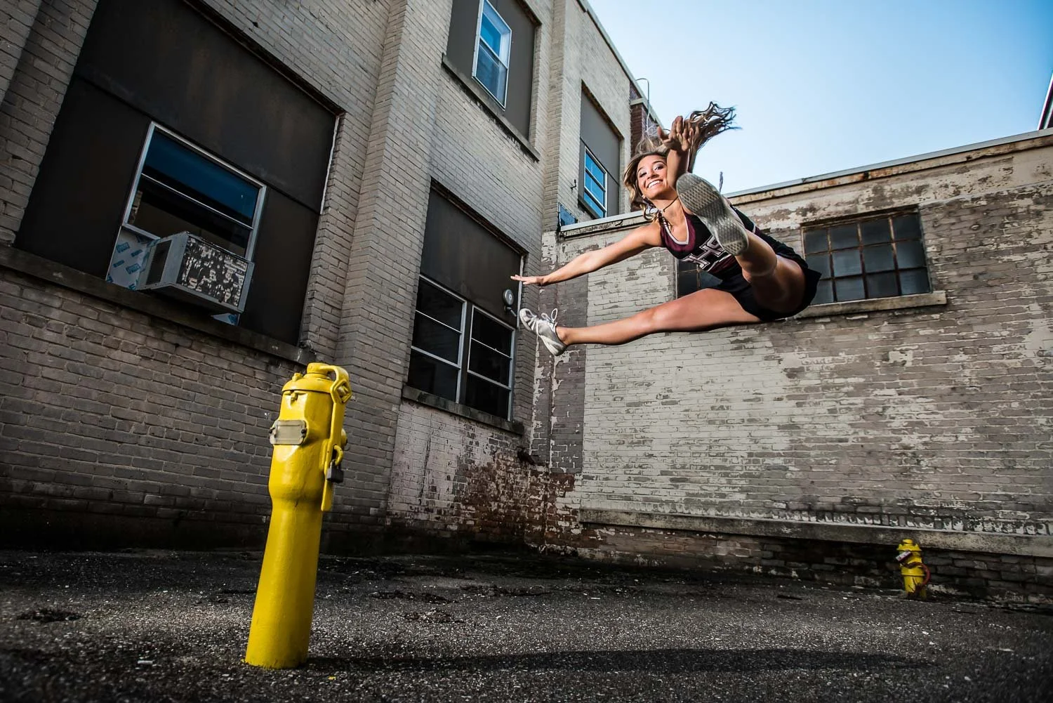 A young woman in athletic attire performing a mid-air split leap between two yellow fire hydrants in an alleyway with brick buildings.