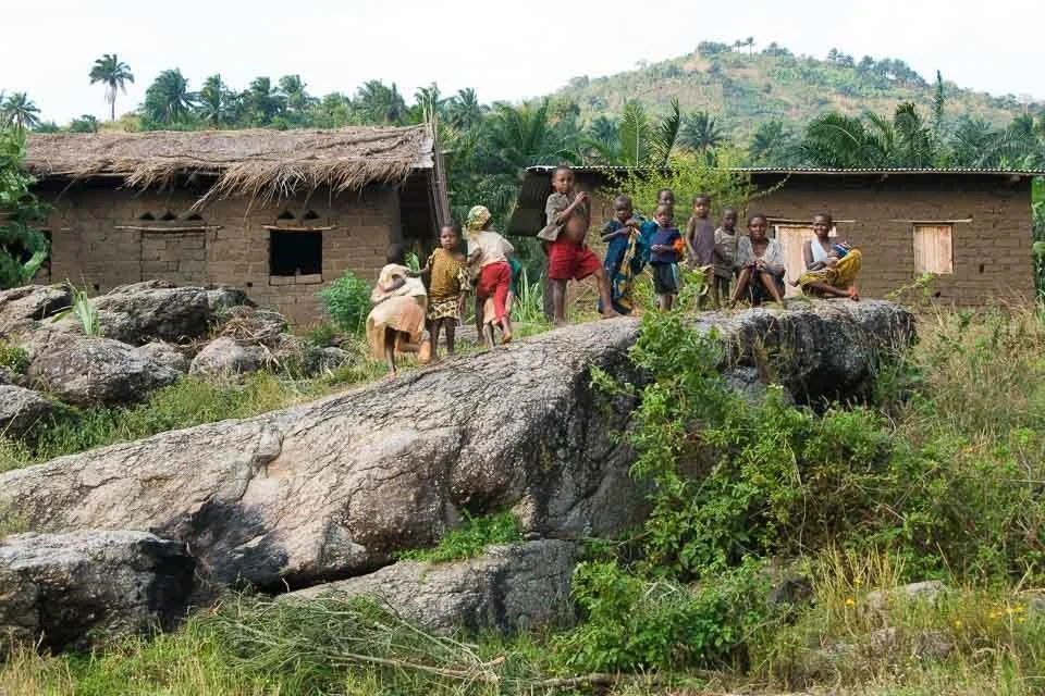Children playing and sitting on rocks in front of mud houses with thatched roofs, surrounded by greenery and hills in the background.
