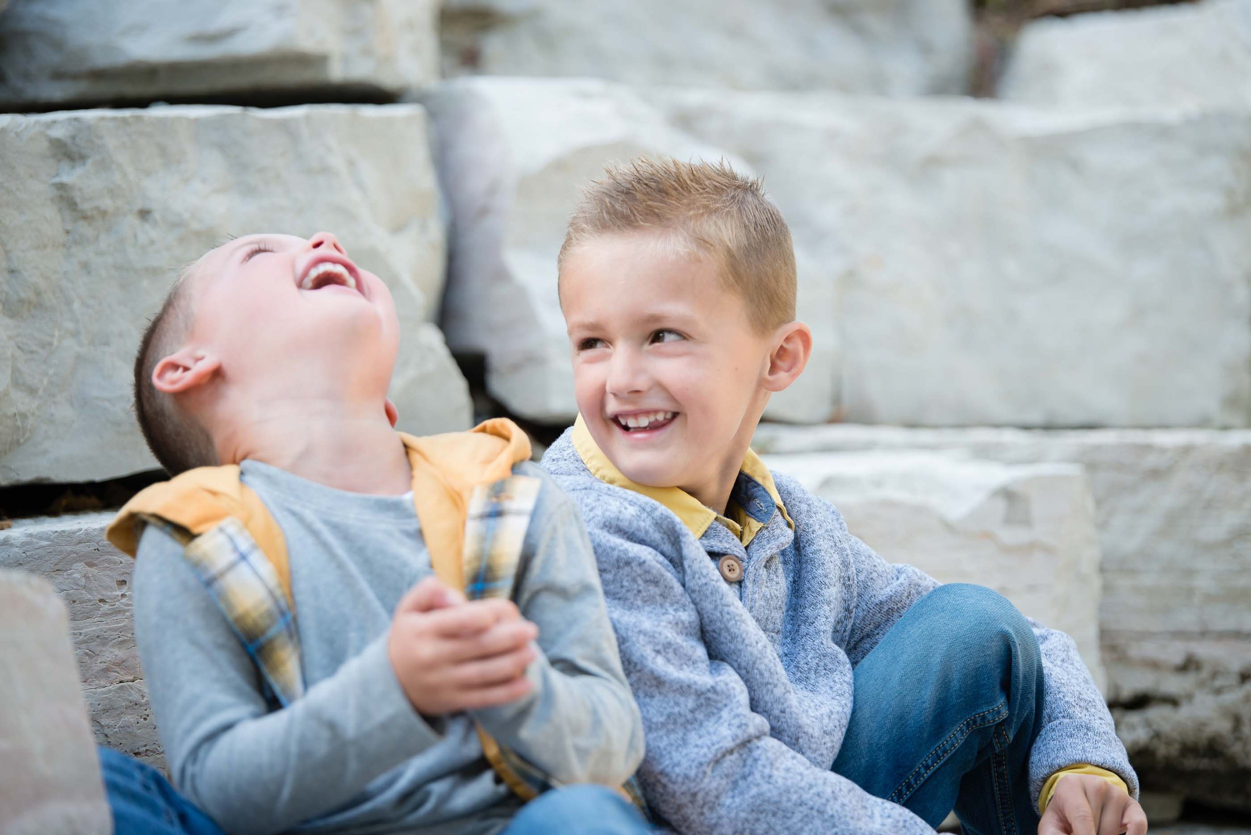 Two young boys sitting outdoors on stone steps, laughing and smiling, wearing casual jackets and jeans.