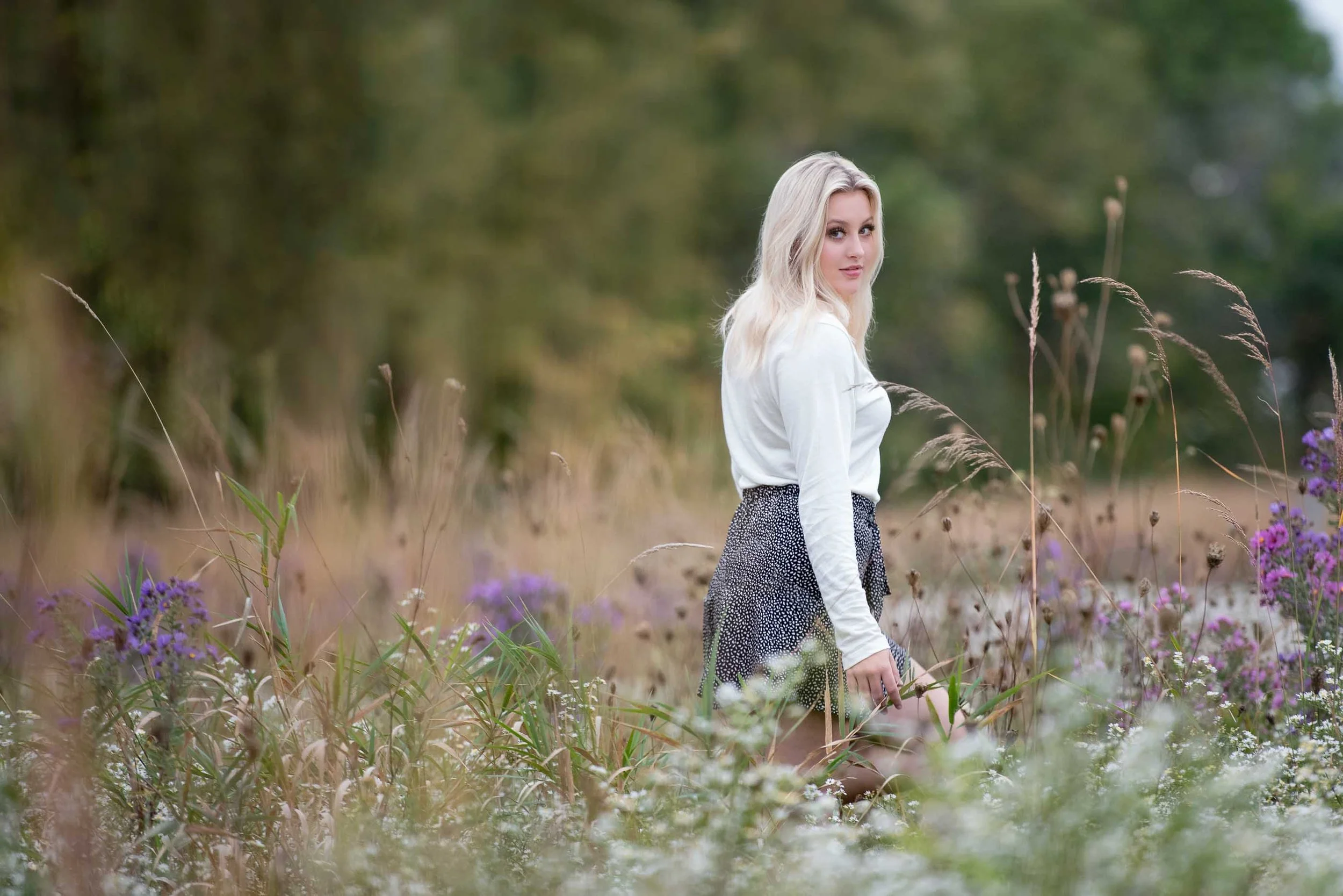 A young blonde woman in a white long-sleeve shirt and a black patterned skirt standing in a field of wildflowers and tall grass with a blurred green forest background.