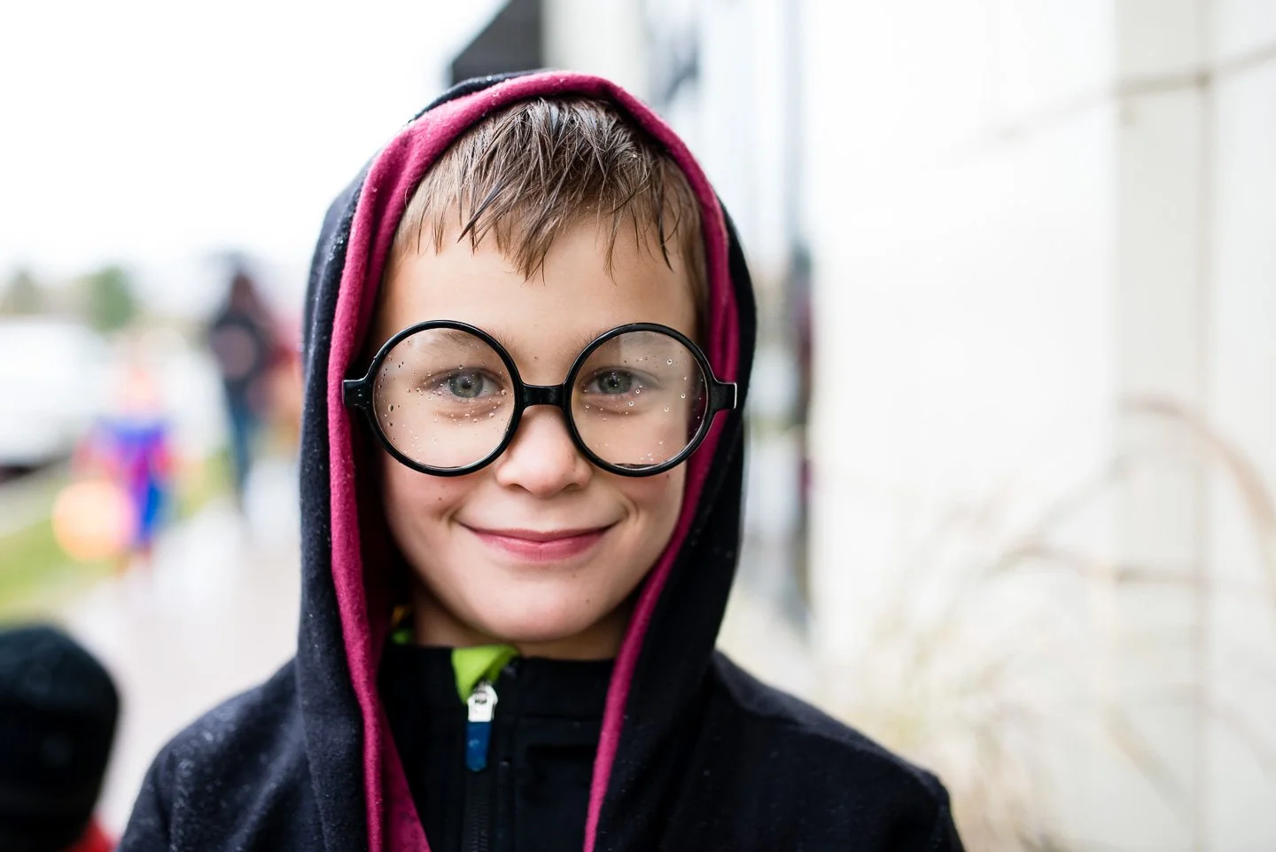 A smiling young boy wearing large black glasses, a black hoodie with a pink lining, and a pink hood, standing outside in the rain.