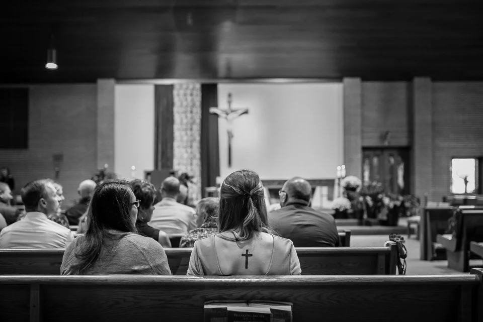 People sitting in pews inside a church, facing the altar with a crucifix hanging on the wall.
