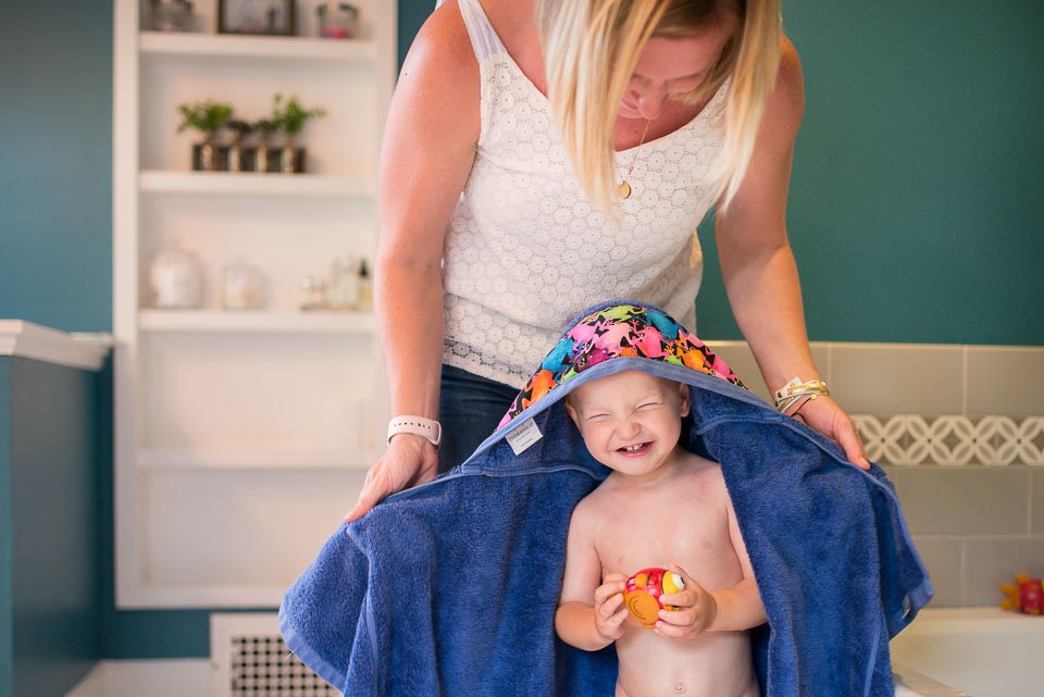 A smiling young boy with a colorful hat and no shirt stands inside a towel tent, holding a rubber duck, while an adult woman, partially visible, leans over him in a bathroom.