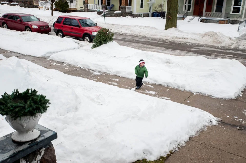 A young child wearing a green jacket and a striped hat walking on a snow-covered sidewalk in a neighborhood with parked cars and colorful houses.