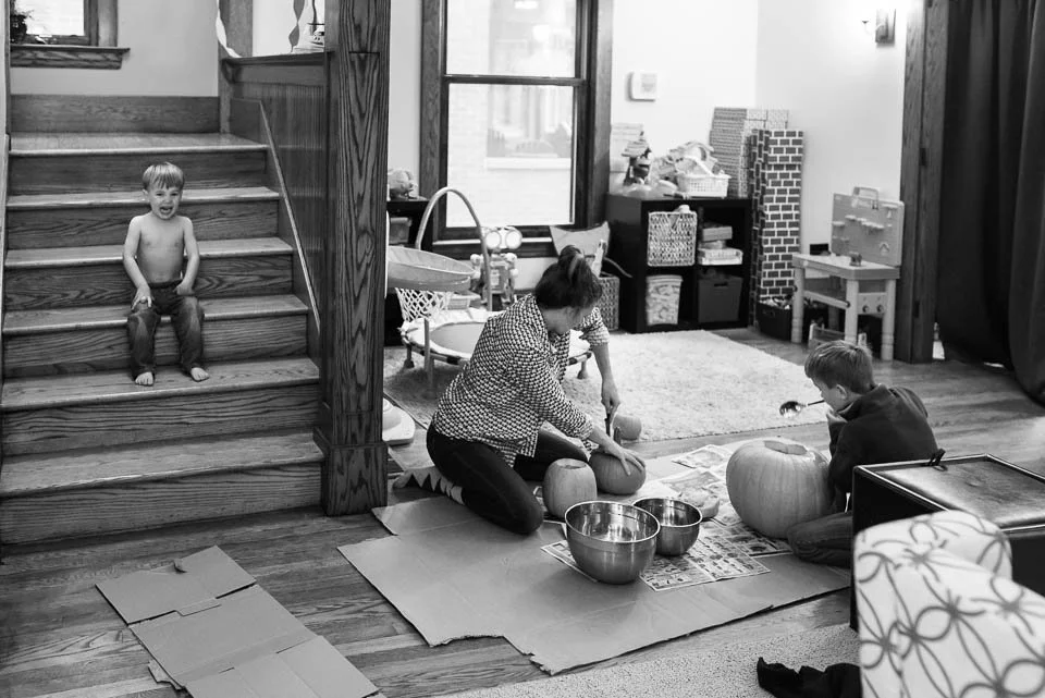 A woman and a child carving pumpkins on a carpeted floor, with a young boy sitting nearby. A boy is sitting on the staircase in the background.