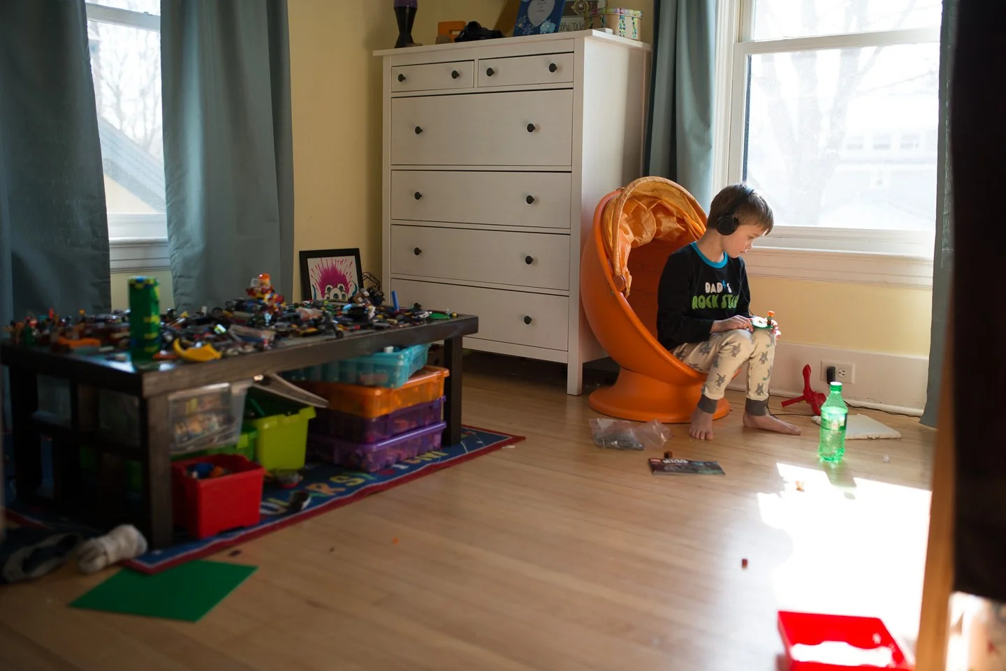 A young boy sitting in an orange egg-shaped chair, wearing headphones and pajamas, playing with toys near a large window in a child's bedroom. There is a white dresser, a table filled with toys, and a green plastic bottle on the floor.