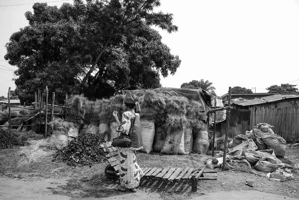 A woman wearing traditional clothing and a head covering stands on a small wooden bridge in front of a large tree, near sacks and bags, in a rural setting.