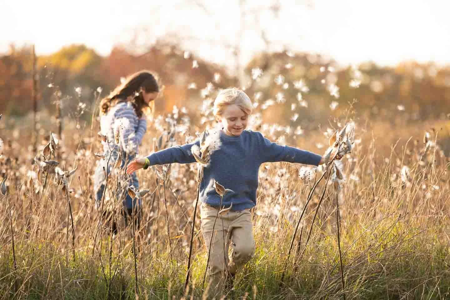 Two children, a boy and a girl, playing in a field of tall grass and dried plants during sunset, with trees in the background.