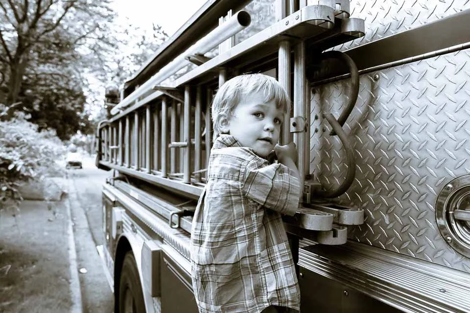 A young boy in a plaid shirt holding onto a fire truck ladder on a park road.