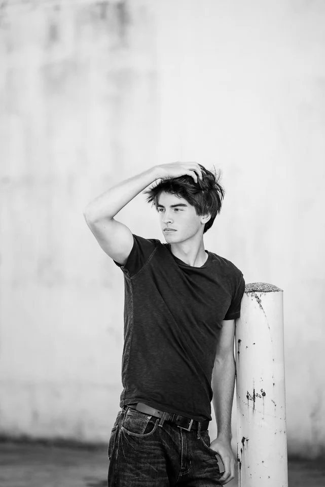 A young man with dark, messy hair and a contemplative expression, standing outdoors in front of a plain wall, wearing a black t-shirt and dark jeans, touching his hair with one hand and leaning on a white bollard.