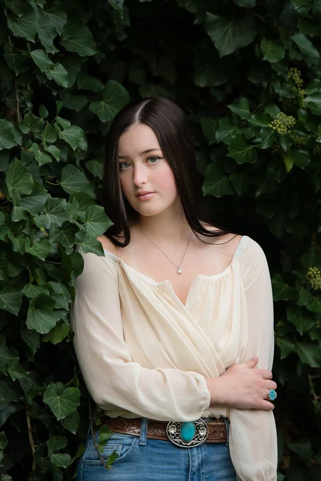 A young woman with long dark hair standing among green ivy leaves, wearing a cream-colored blouse, blue jeans, a turquoise ring, and a necklace.