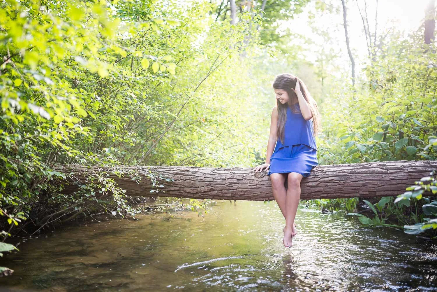 A young woman in a blue dress sitting on a fallen tree trunk over a clear stream in a lush green forest, smiling and touching her hair.