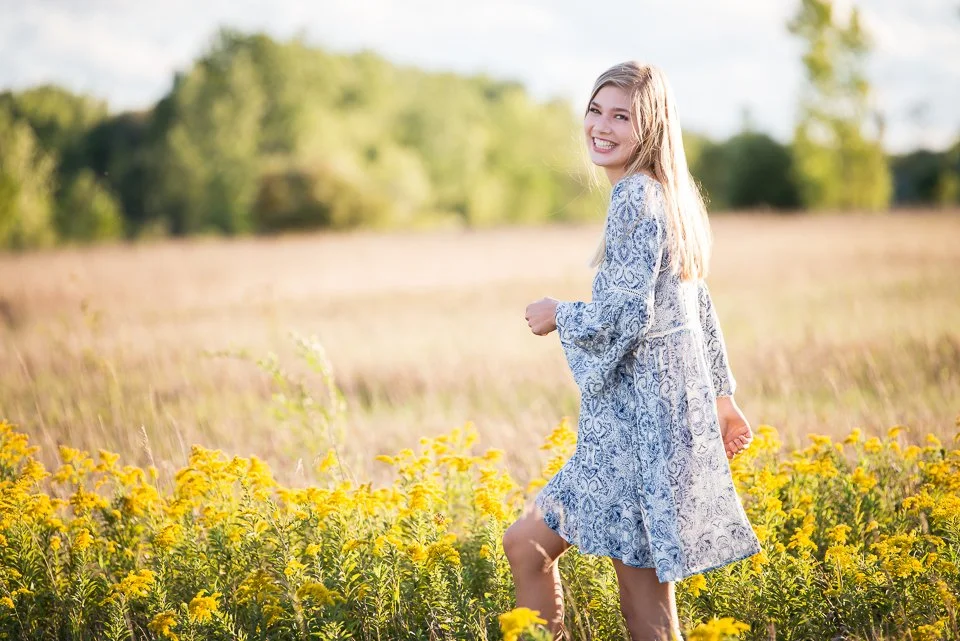 A young woman with long blonde hair smiling and wearing a light blue patterned dress standing in a field of yellow flowers on a sunny day.