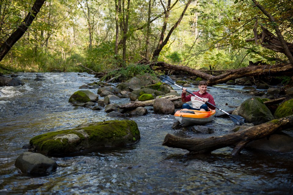 A boy kayaking on a rocky stream in a forested area.