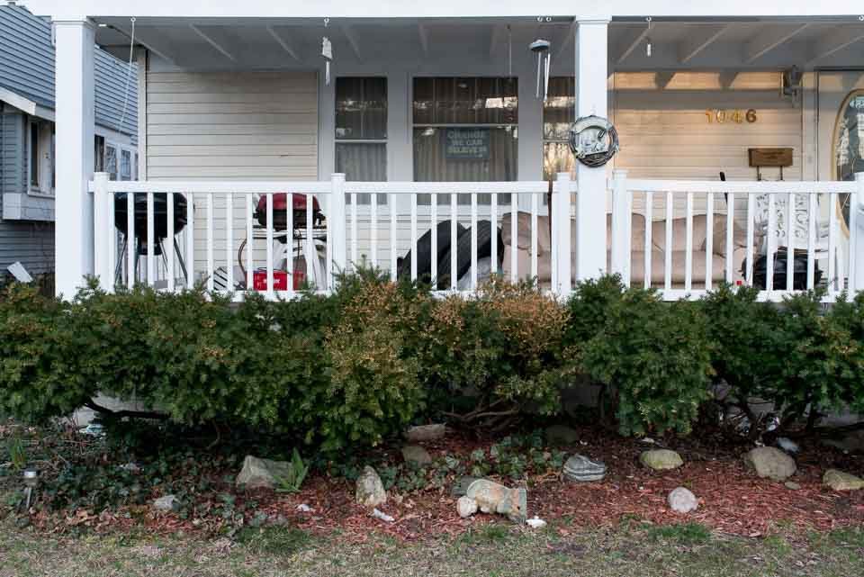 Front porch of a house with white railing, two chairs, a red cooler, a grill, and a hanging helmet. Bushes with rocks in front and house number 1046 displayed.