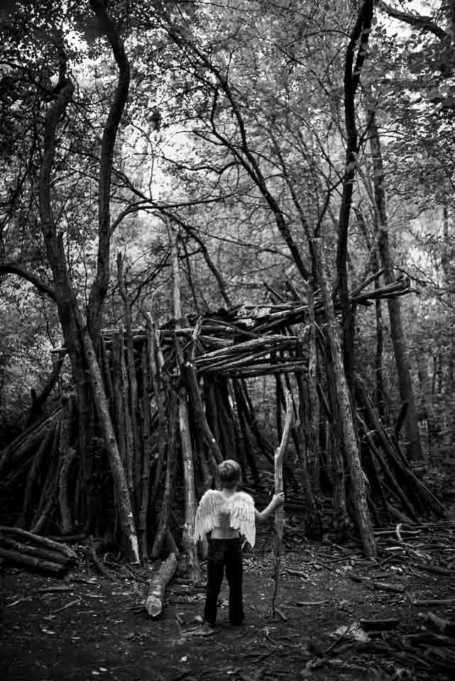 A young girl dressed as an angel with wings and holding a staff stands in front of a large, makeshift wooden shelter in a wooded area, in black and white.