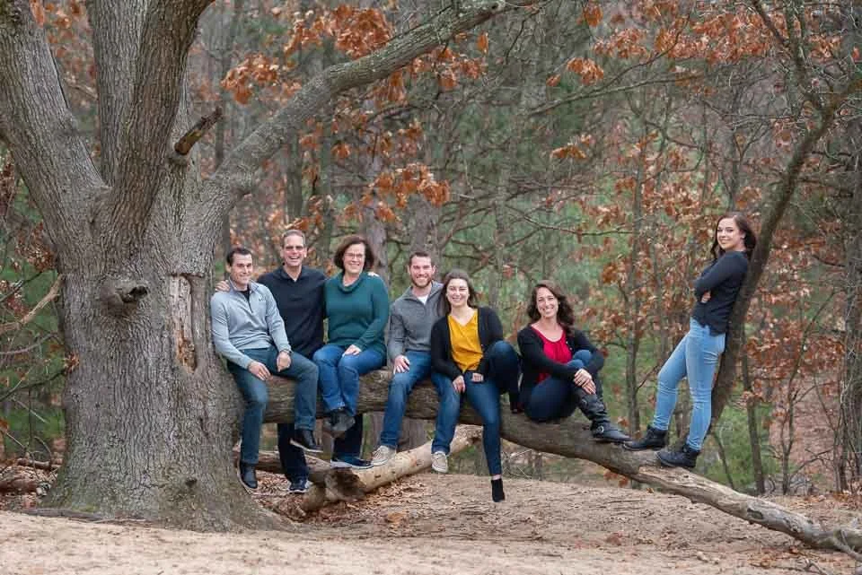 A group of seven people sitting and standing on a large tree branch in a wooded area during fall.