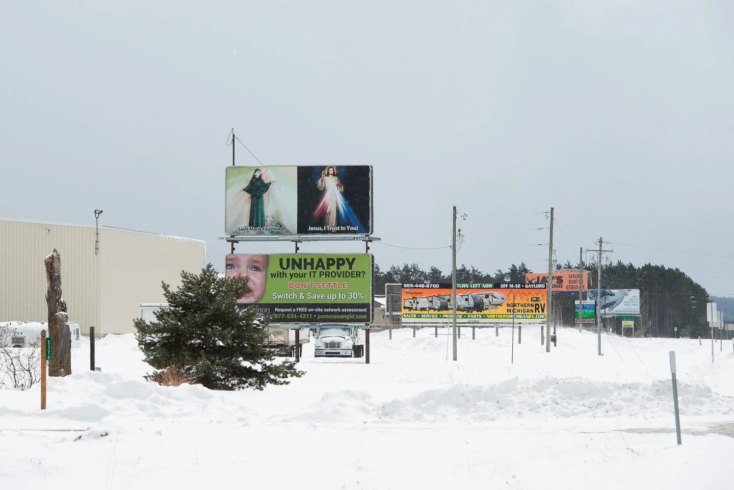 Snow-covered street with multiple billboards and power lines, featuring advertisements for religious images, an IT service provider, and a Recreational Vehicle dealership.