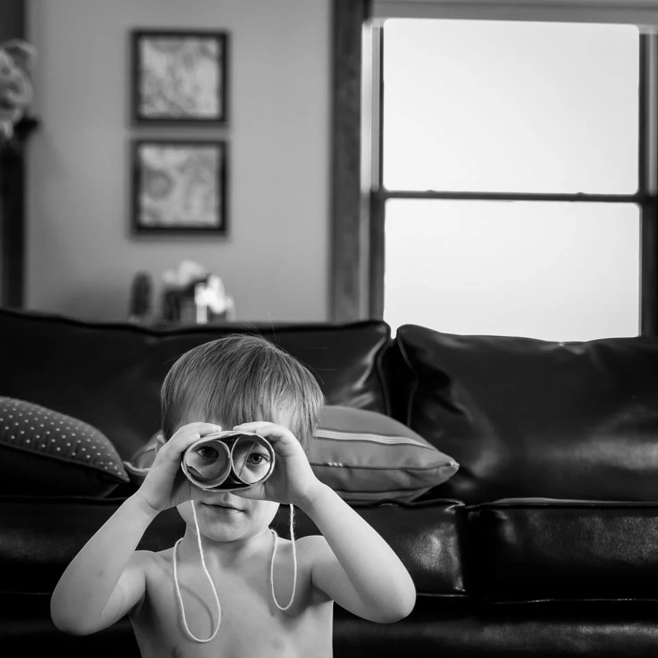 A young boy is sitting on a couch, holding a pair of cardboard tubes like binoculars to his eyes, in a living room with a window and framed pictures on the wall.