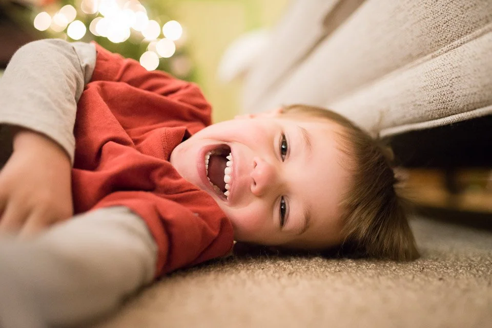Young boy lying on the floor, smiling and laughing, with Christmas lights blurred in the background.