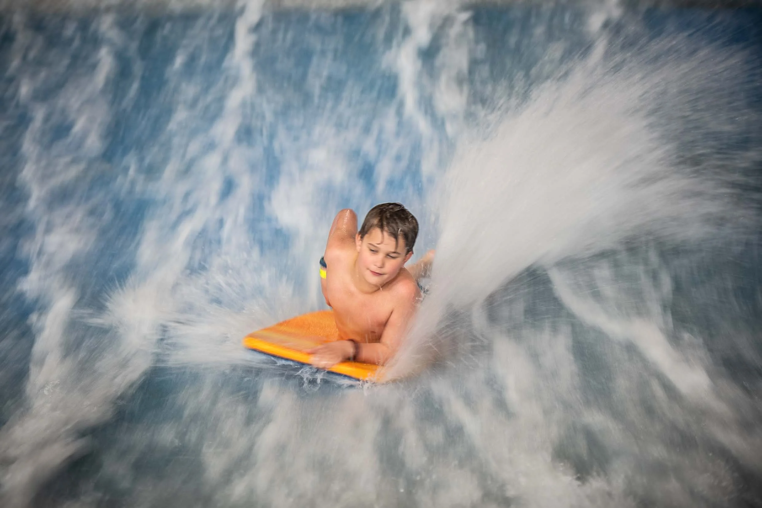 A young boy wearing swim trunks riding a wave on an orange boogie board in the ocean.