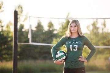 A young woman in a green volleyball uniform holding a volleyball on an outdoor volleyball court.