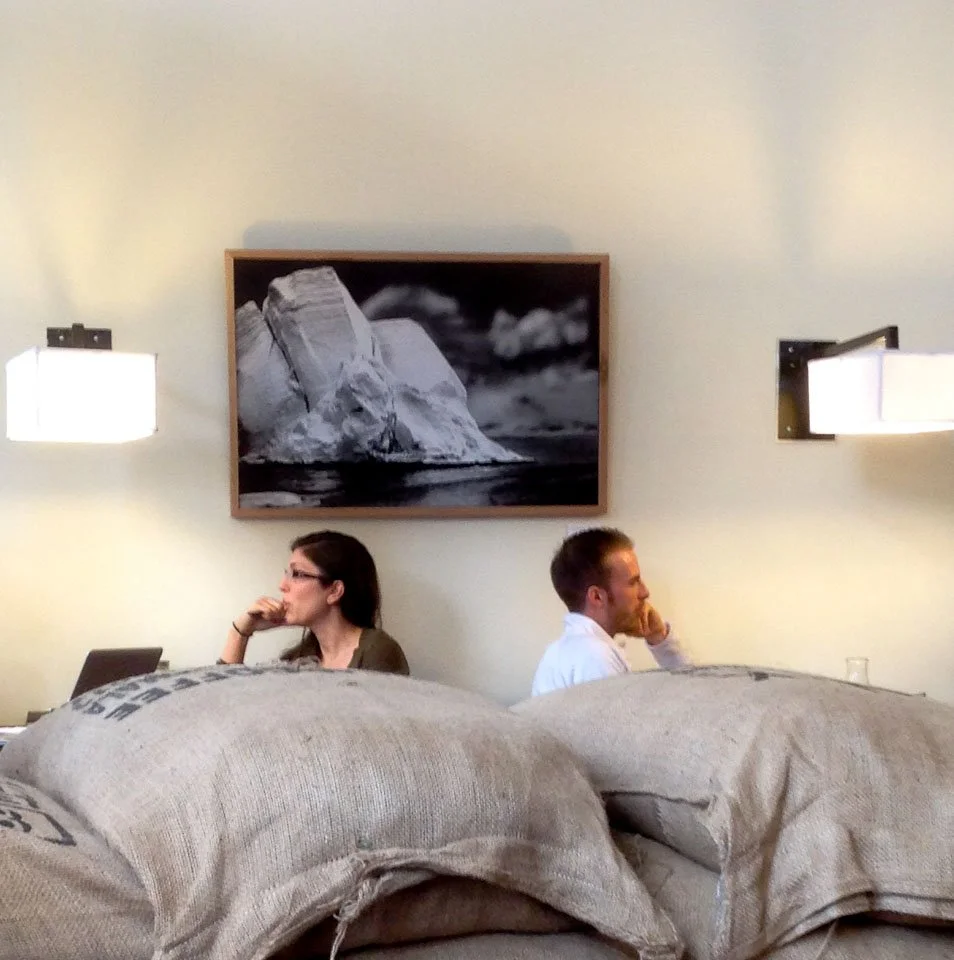 Two people sitting back-to-back on beds in a hotel room, each using a laptop, with a framed black-and-white photo hanging on the wall above them.