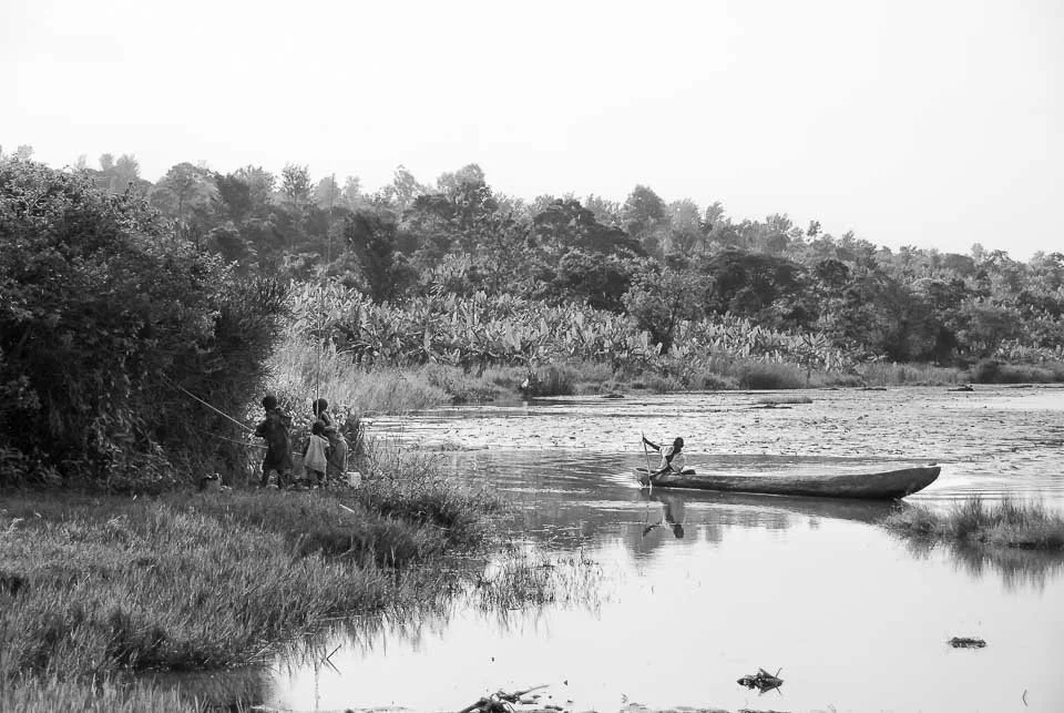 People fishing near a lake with a boat, surrounded by trees and vegetation in black and white.