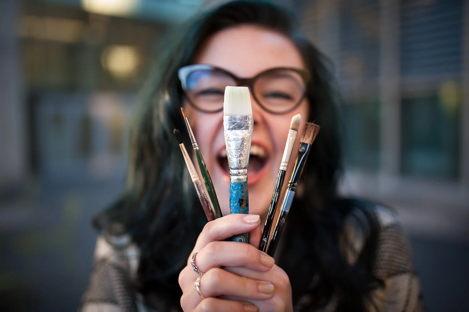 A woman with glasses smiling and holding several paintbrushes close to her face outdoors.