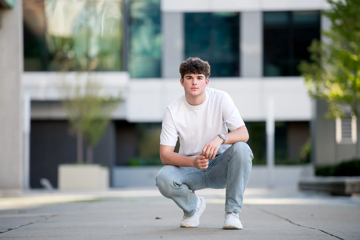 A young man squatting outdoors in front of a modern glass building, wearing a white t-shirt, gray jeans, and white sneakers.