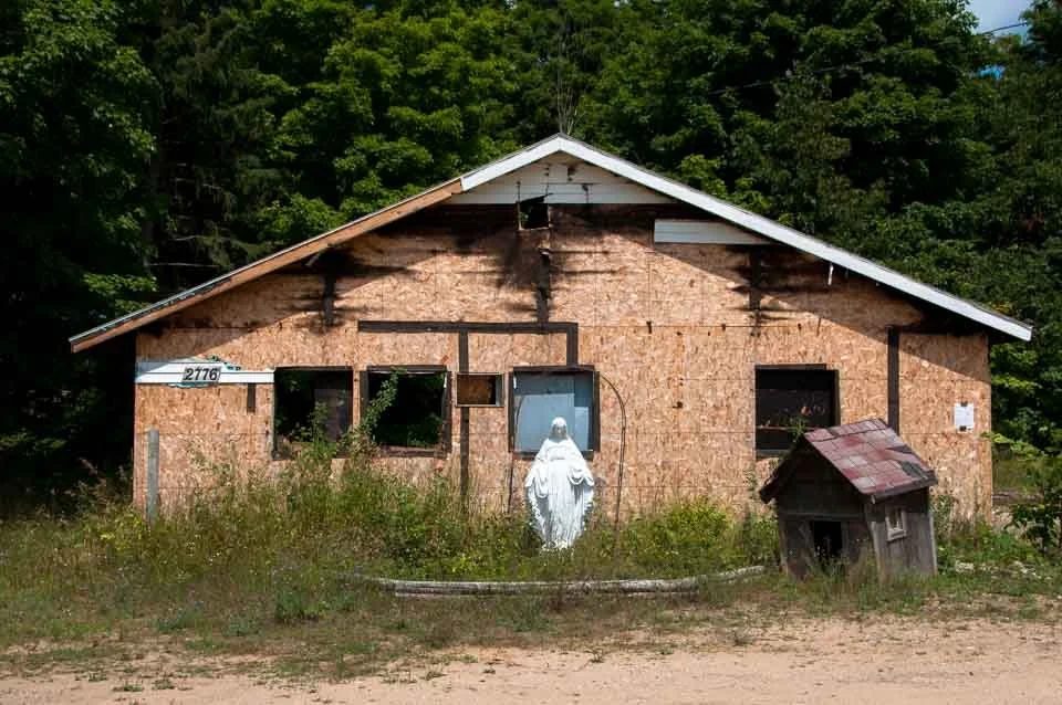 Unfinished house with plywood walls, a cross on the front, and a statue of the Virgin Mary in front, surrounded by grass and bordered by trees.