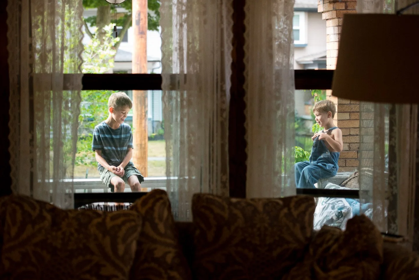 Two boys sitting and smiling on a porch outside a house, viewed through a window with lace curtains inside a living room.