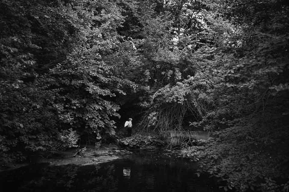 Person standing on a rocky bank by a stream surrounded by dense trees and foliage.