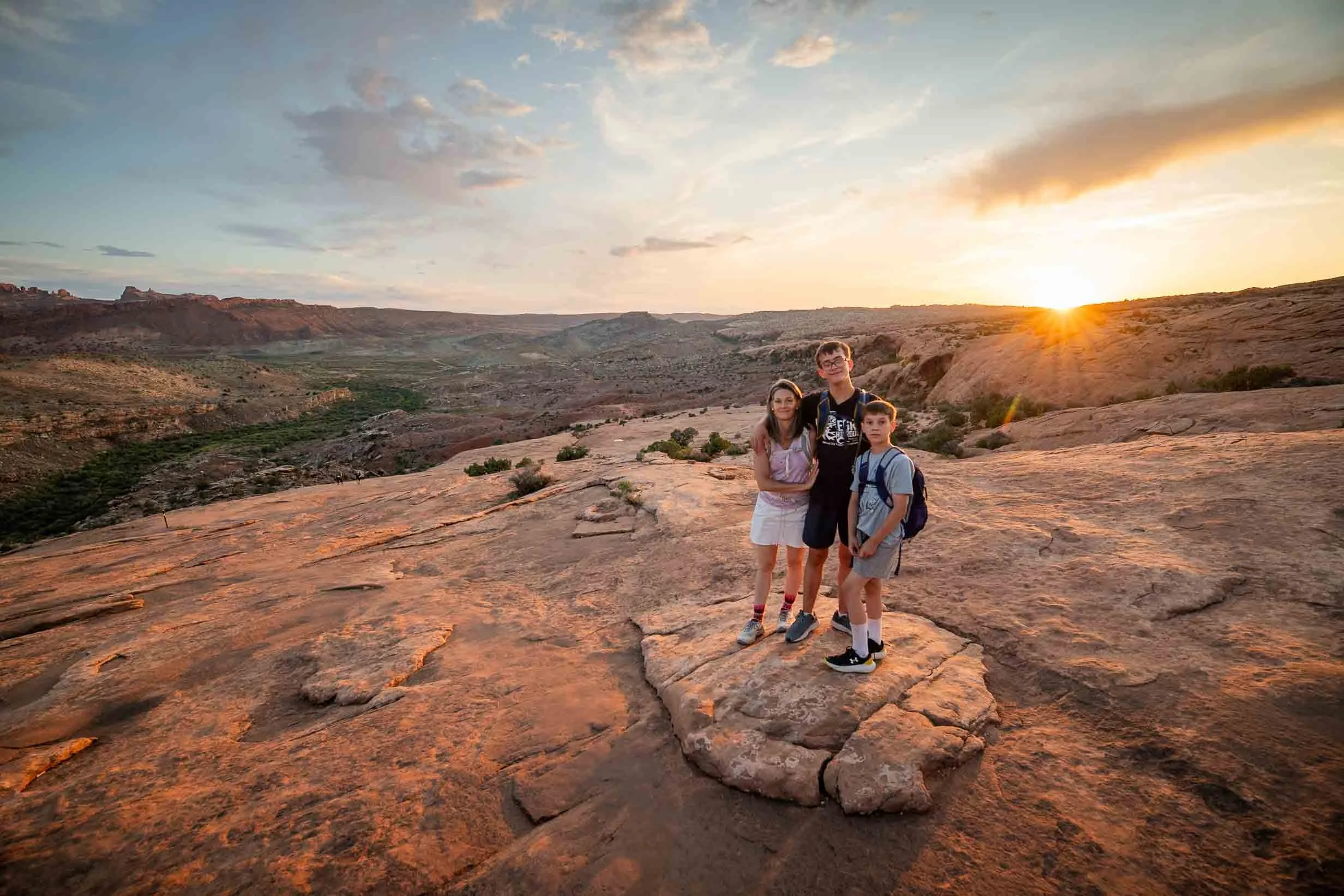 Three children standing on a large rock during sunset in a desert landscape with cliffs and sparse vegetation in the background.