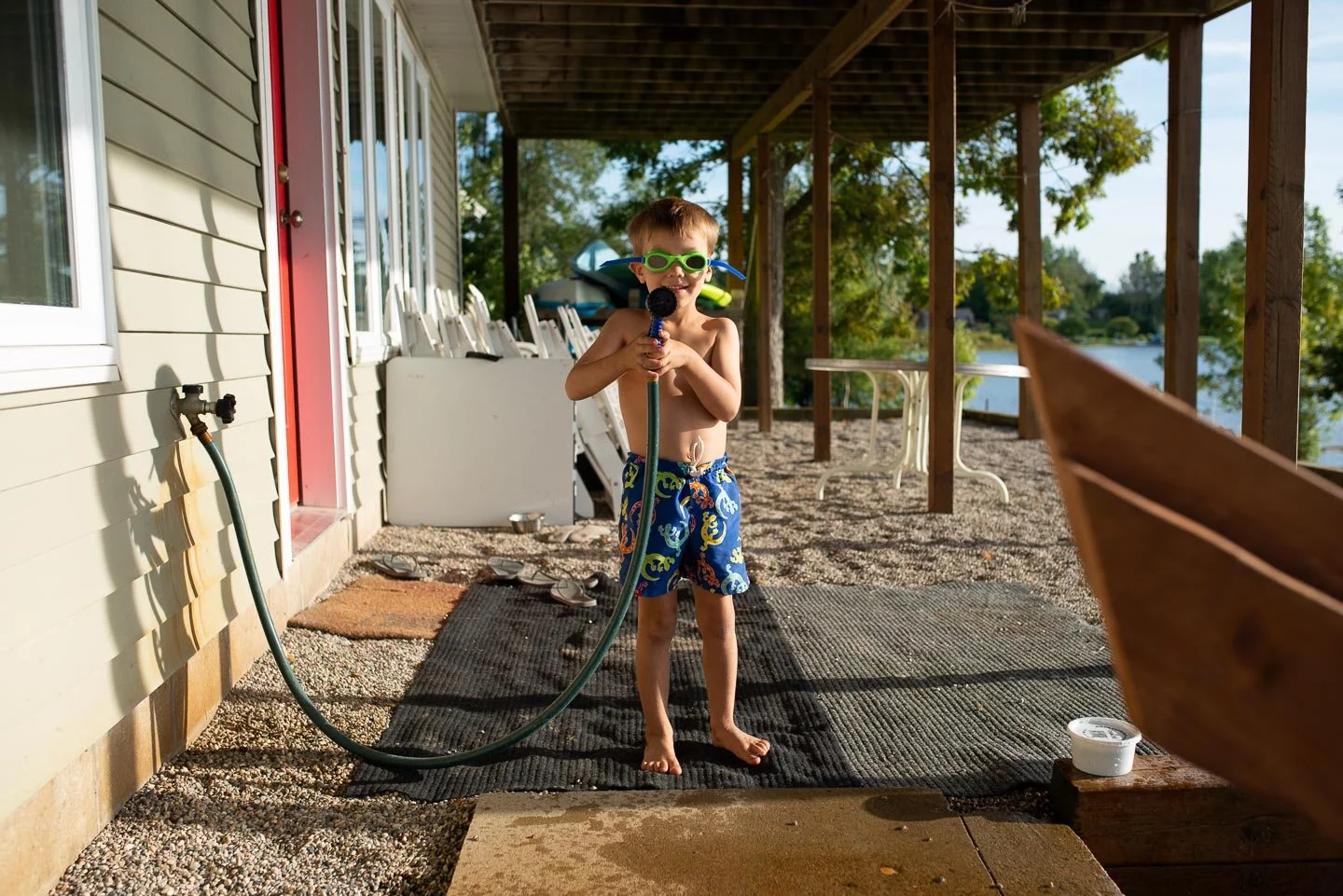 A young boy in swim trunks and goggles standing outdoors on a gravel porch, holding a water hose and smiling.