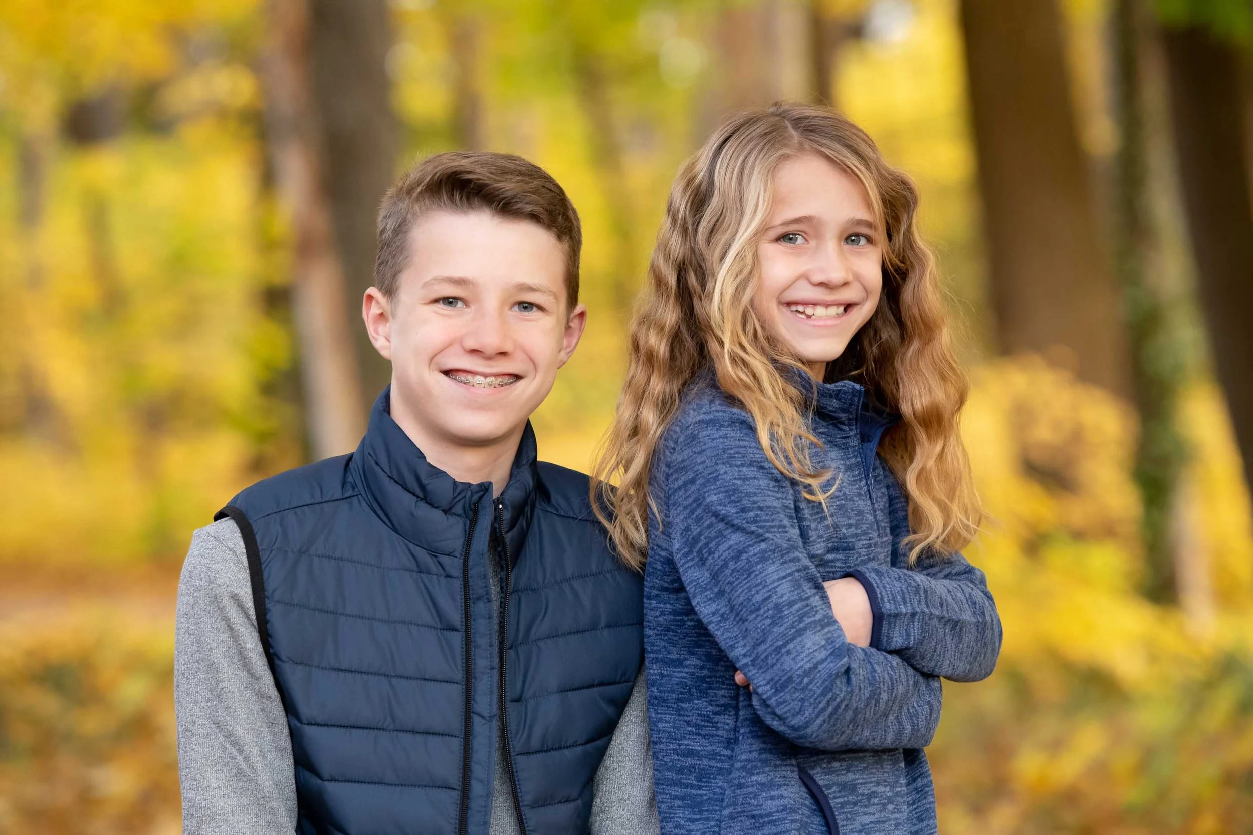 Two kids, a boy and a girl, standing in a forest with autumn foliage, smiling at the camera.