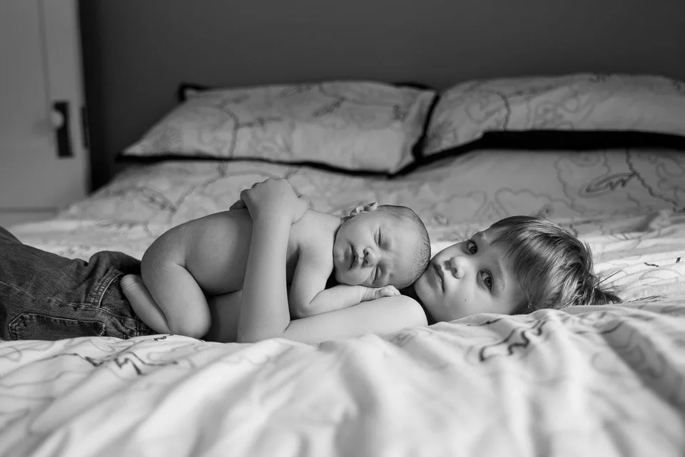 A young girl lying on a bed, holding a sleeping newborn baby on her chest, in a monochrome photo.