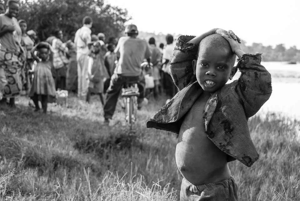 A young boy standing in a field, holding his head with both hands, with a group of people in the background.