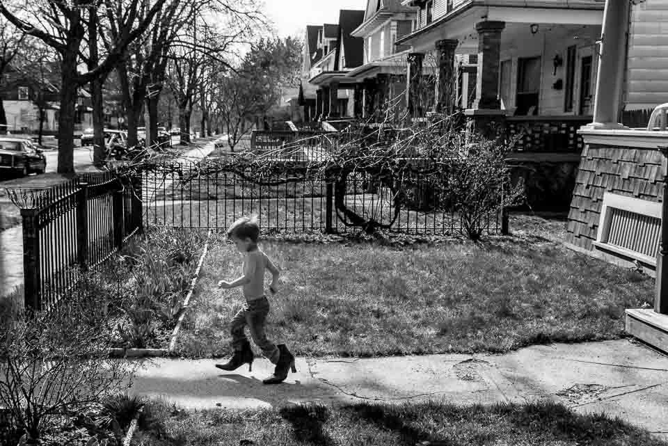A young boy walking on a sidewalk in a small yard with grass, surrounded by a metal fence, in front of a row of houses with porches and trees in the background, black and white photo.