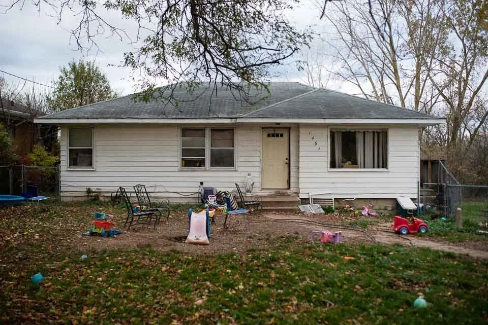 A white house with a gray roof and front stairs, surrounded by leafless trees, with toys scattered in the yard.