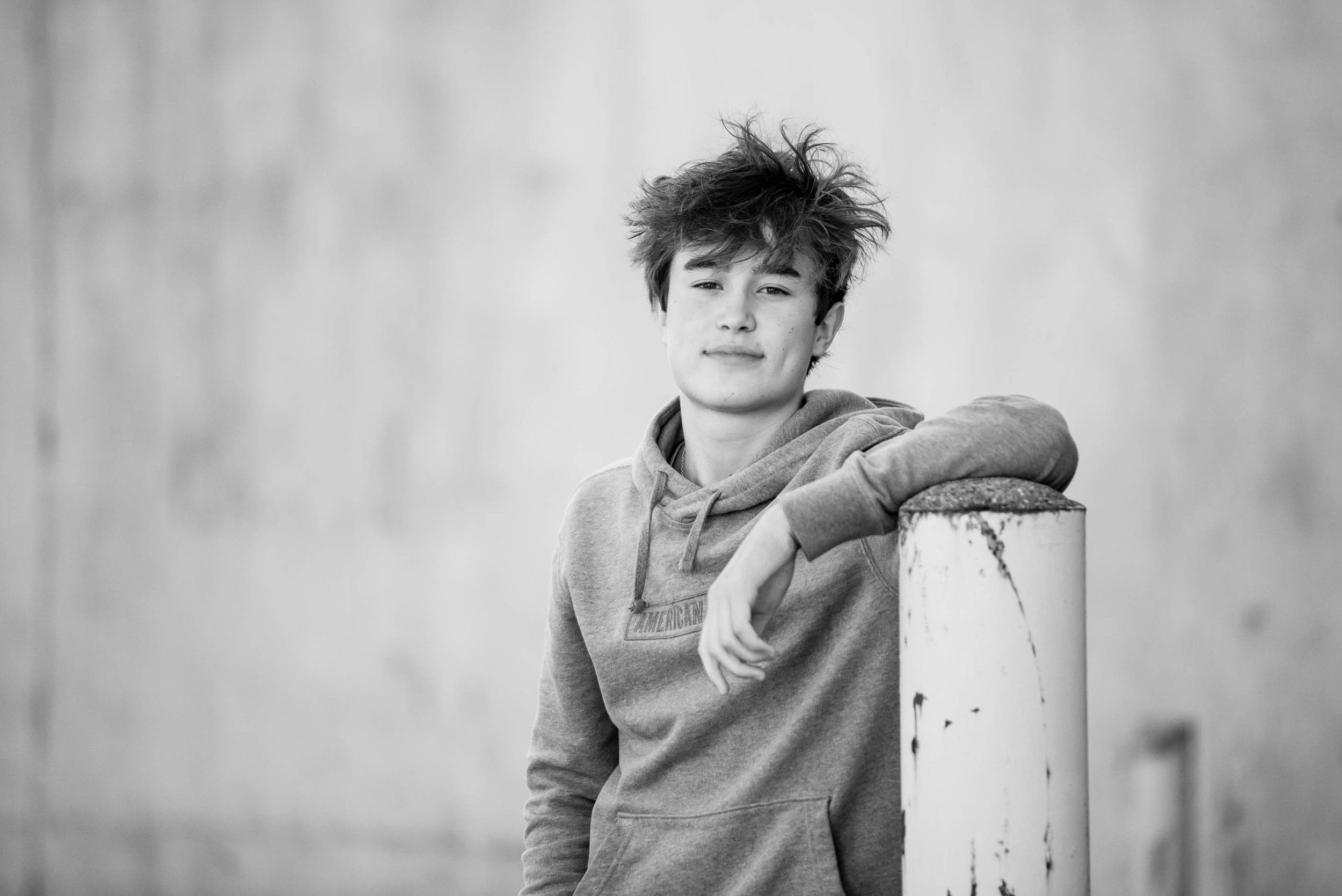 Black and white photo of a teenage boy with messy hair leaning on a post, wearing a hoodie, looking at the camera with a slight smile.