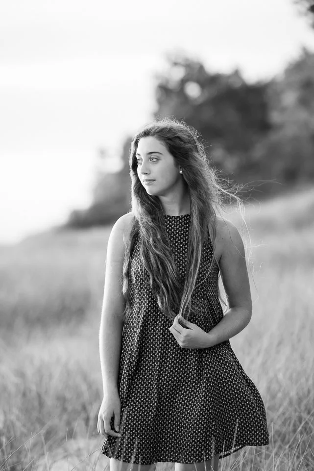 Black and white photo of a young woman with long hair standing outdoors in a grassy field with trees in the background, looking to her left.