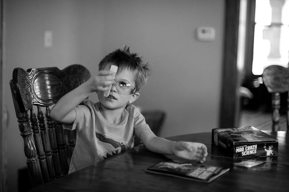 A young boy sitting at a dining table holding a small object up to his eye, wearing safety glasses. There is a science kit box labeled 'Mad Crazy Science' and a booklet on the table in front of him.