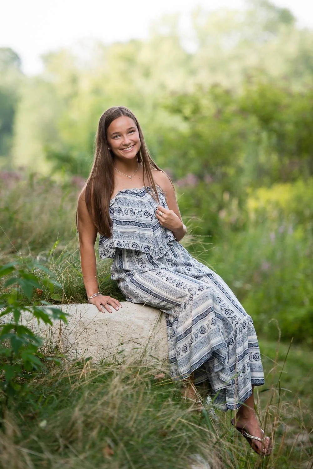 A young woman with long brown hair sitting on a large rock outdoors in a natural setting surrounded by green foliage, smiling at the camera, wearing a patterned sleeveless dress and sandals.
