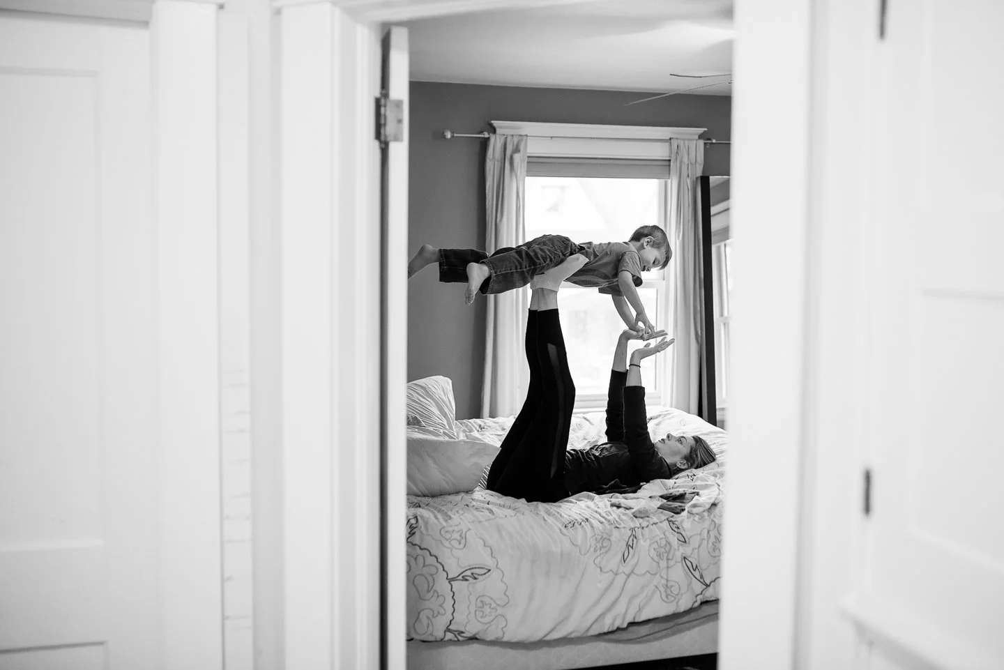 A woman lying on a bed holding a young boy in the air, who is balancing on her hands and reaching towards another child, in a bedroom with a window and curtains.