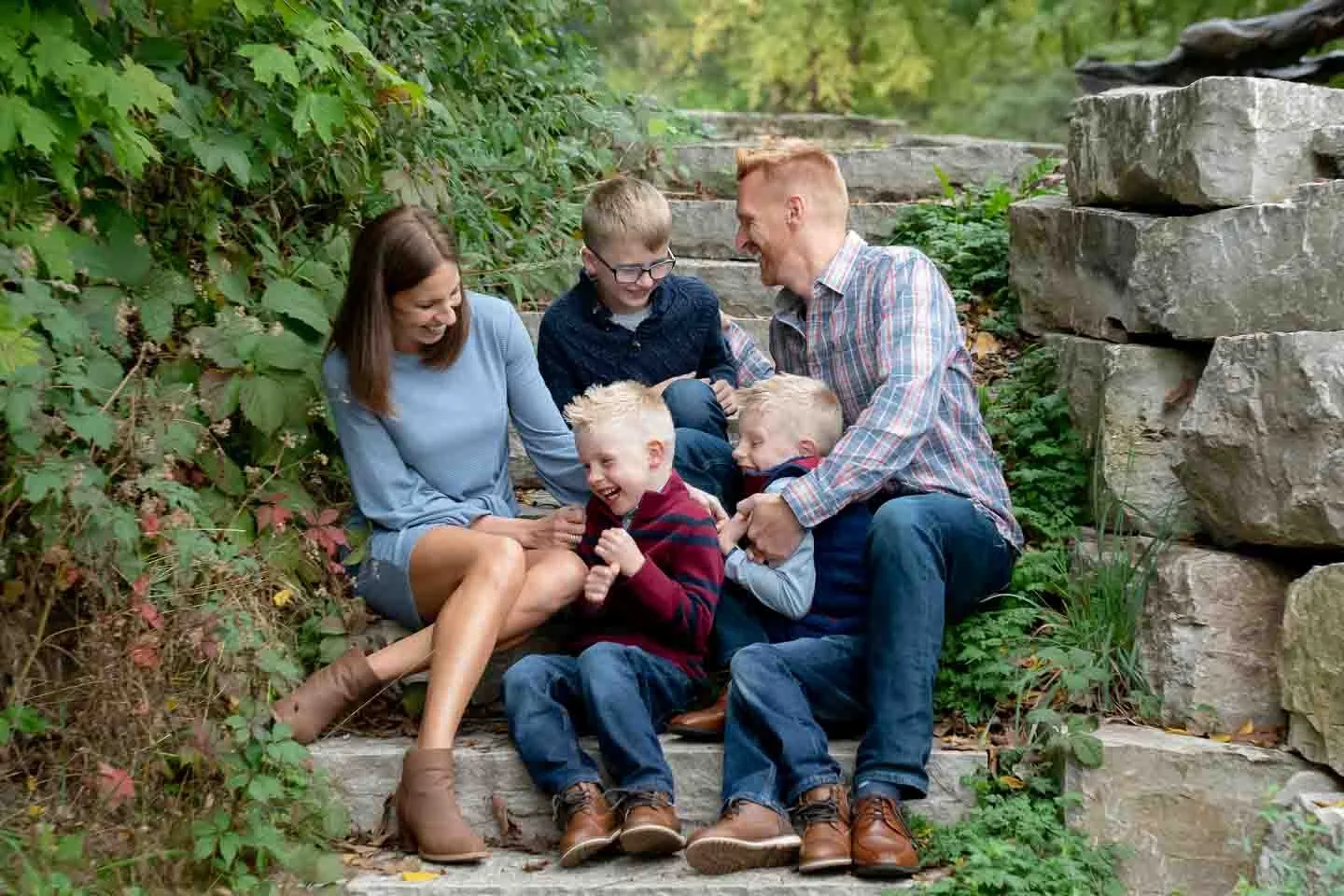 A family of six sitting on outdoor stone steps in a green, leafy park, laughing and playing together.