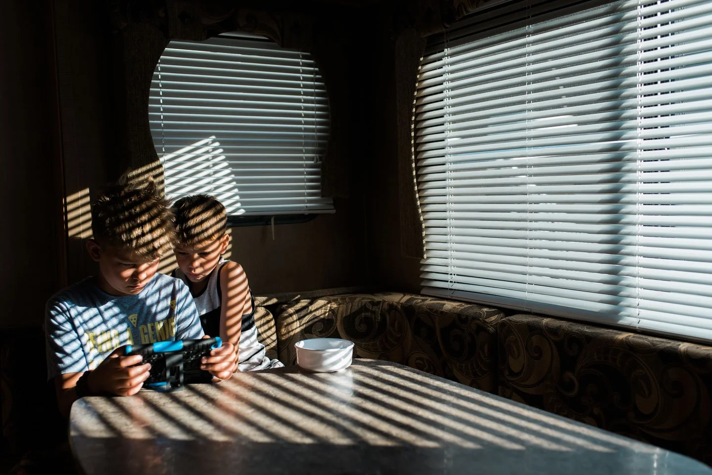 Two young boys sit at a table in a room with closed blinds, sunlight streaming through creating striped shadows on their faces, table, and wall.