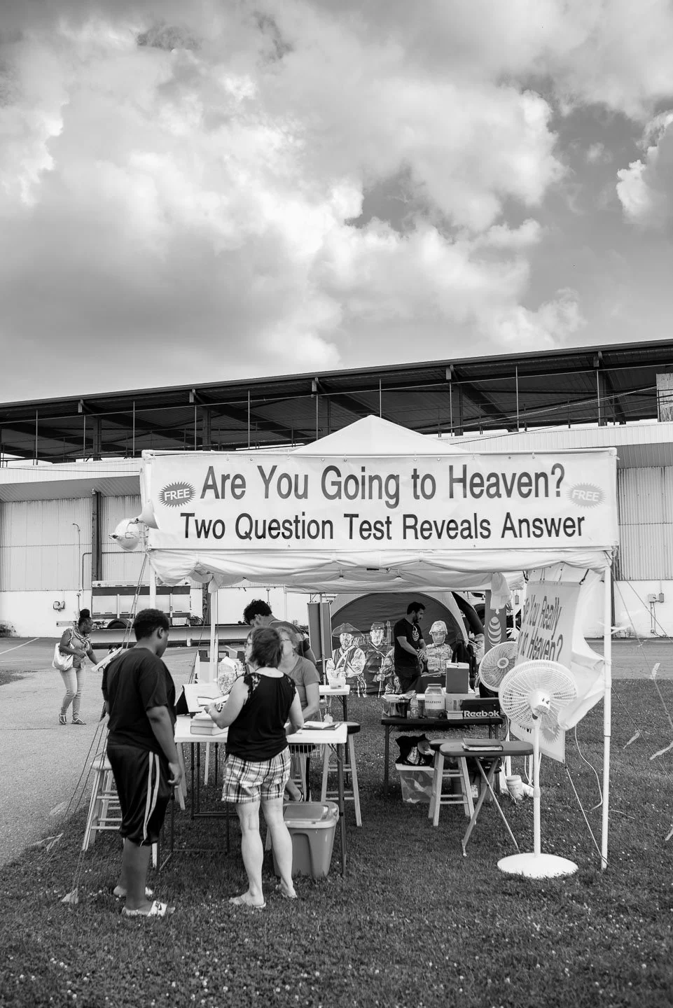 A booth with a sign that reads 'Are You Going to Heaven? Two Question Test Reveals Answer,' at an outdoor event, with people gathered around, some reading materials, and pollinating fans on the side.