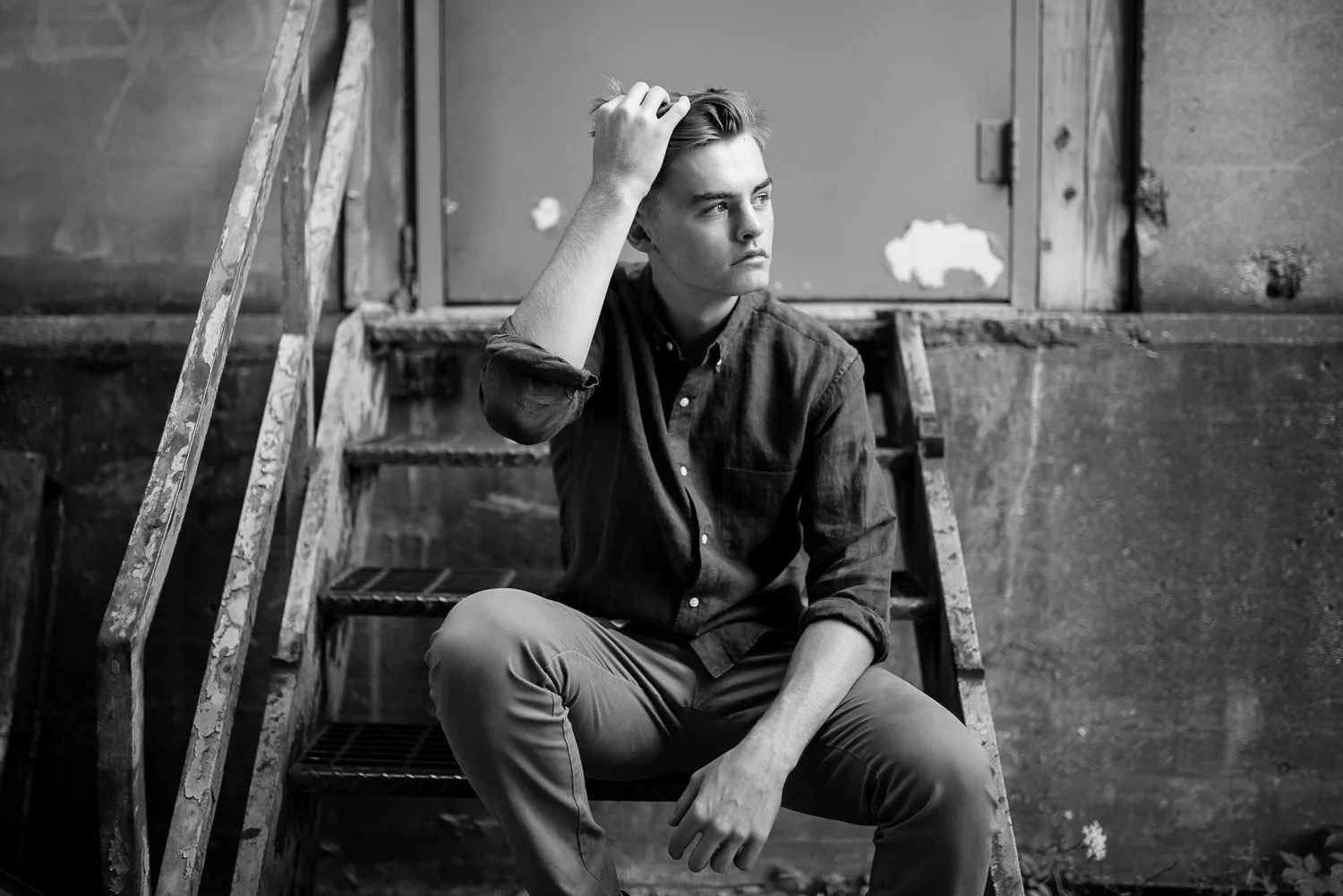A young man with light-colored hair sitting on stairs, scratching his head and looking thoughtful, in an old building.
